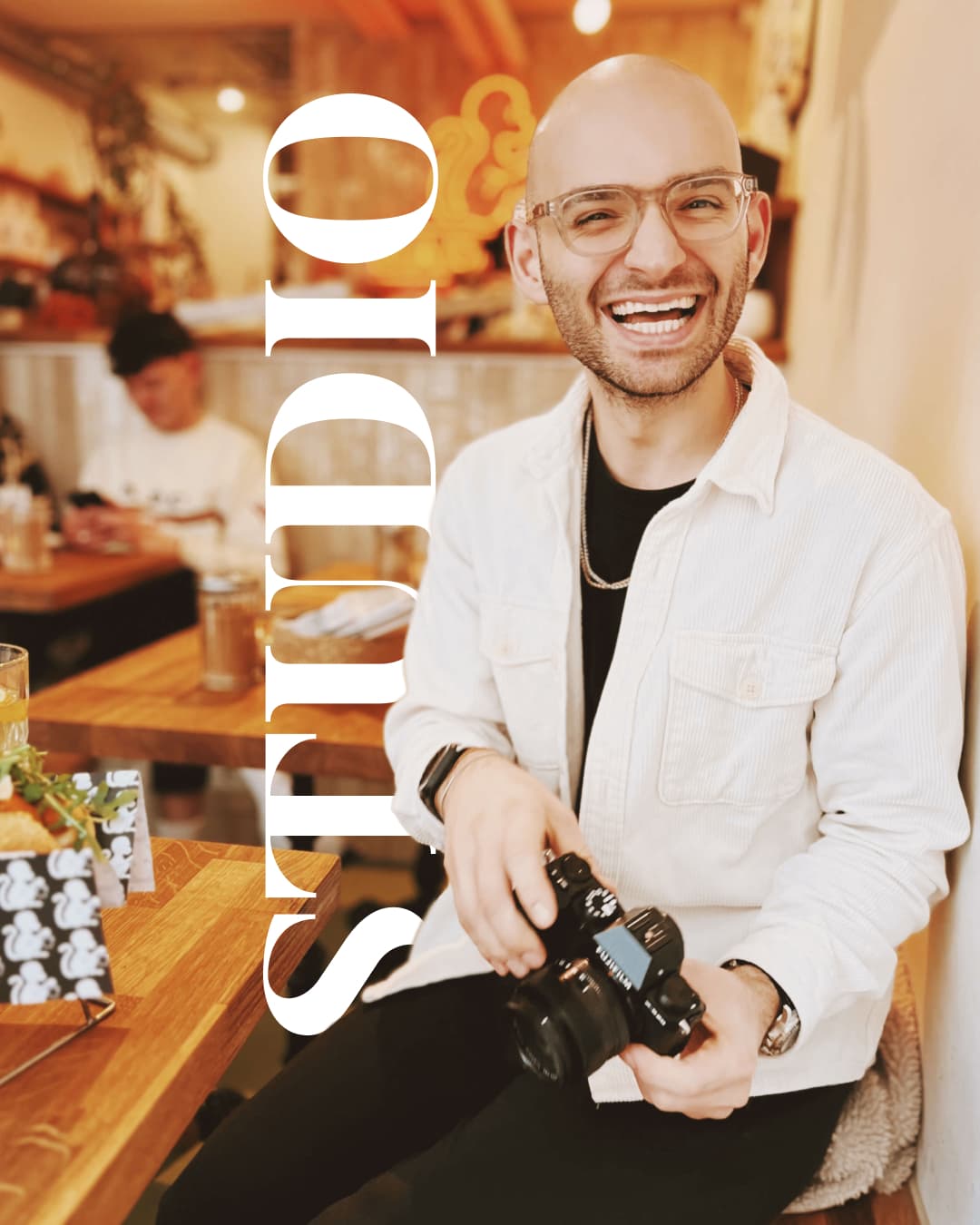 Lachender Designer mit Kamera in Café. "STUDIO"-Schriftzug symbolisiert Selbstvertrauen und Freude an kreativer Selbstständigkeit.