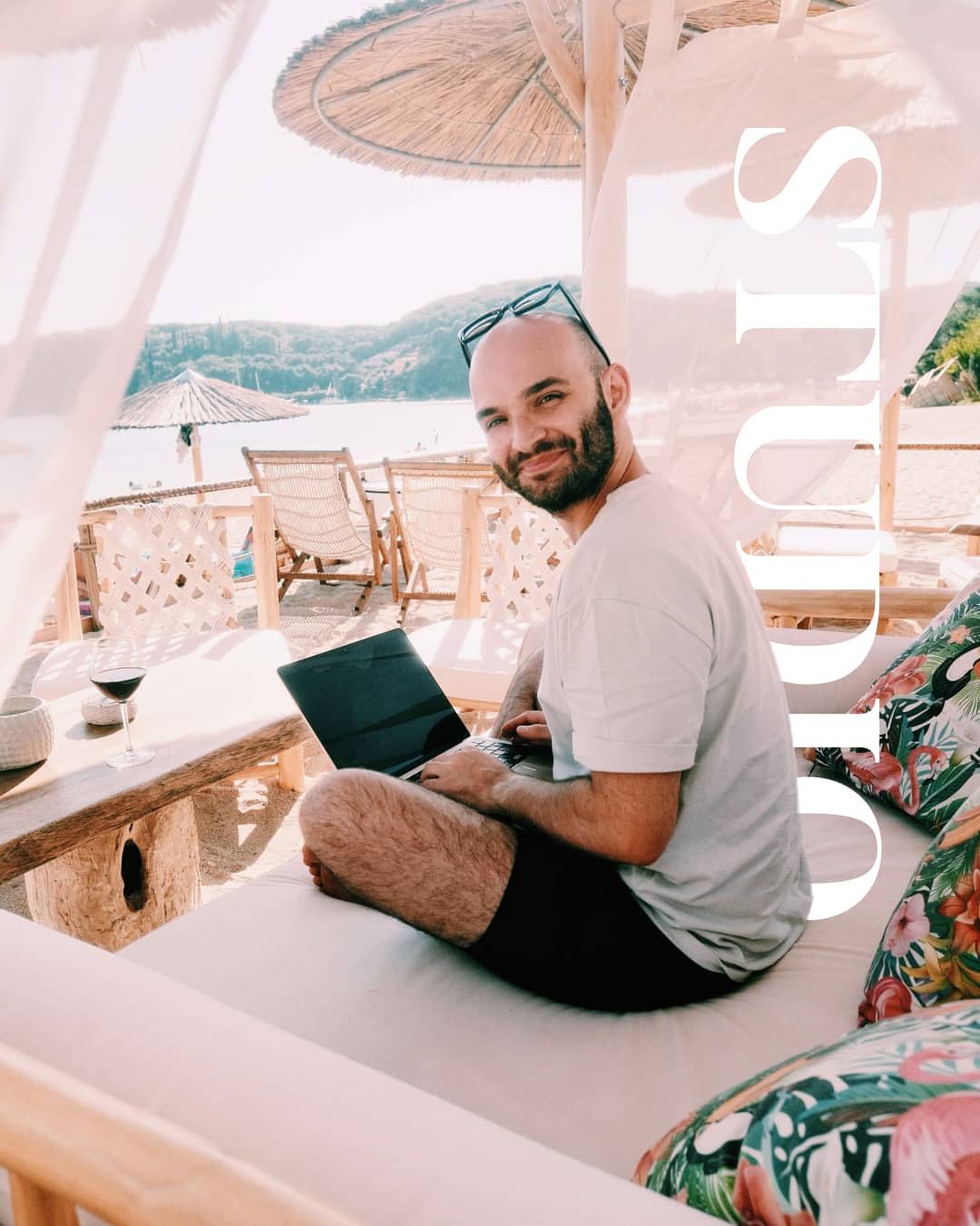 Digital Nomad arbeitet entspannt mit Laptop am Strand. Sonnenschirm, Meer und Berge im Hintergrund. Authentisches Personal Branding in Aktion.