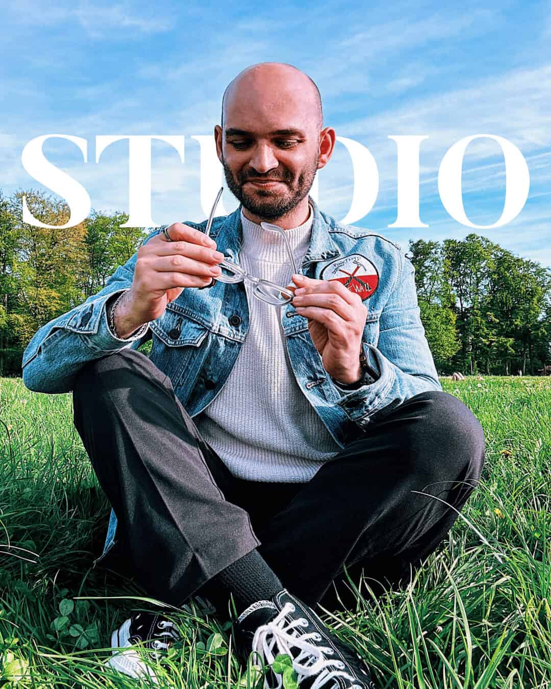 Mann sitzt entspannt im Gras, betrachtet Brille. Symbolisiert Geduld und Reflexion für Selbstständige. STUDIO-Schriftzug im Hintergrund.