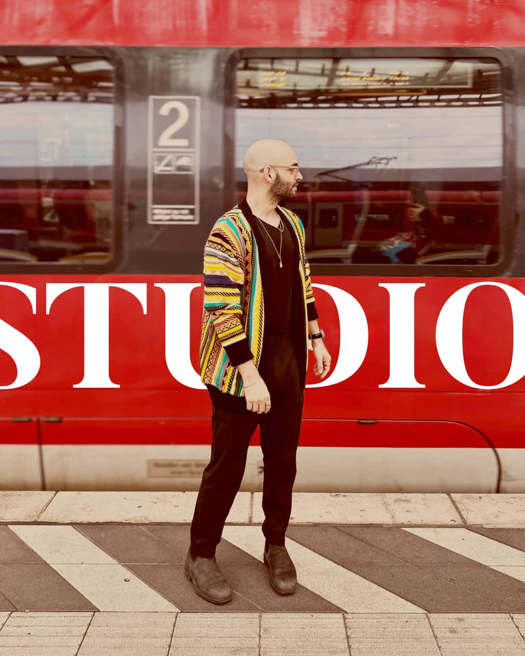 Kreativer UX-Designer mit bunter Jacke vor rotem Zug am Bahnsteig. "STUDIO" sichtbar, symbolisiert Selbstständigkeit und Design-Expertise.