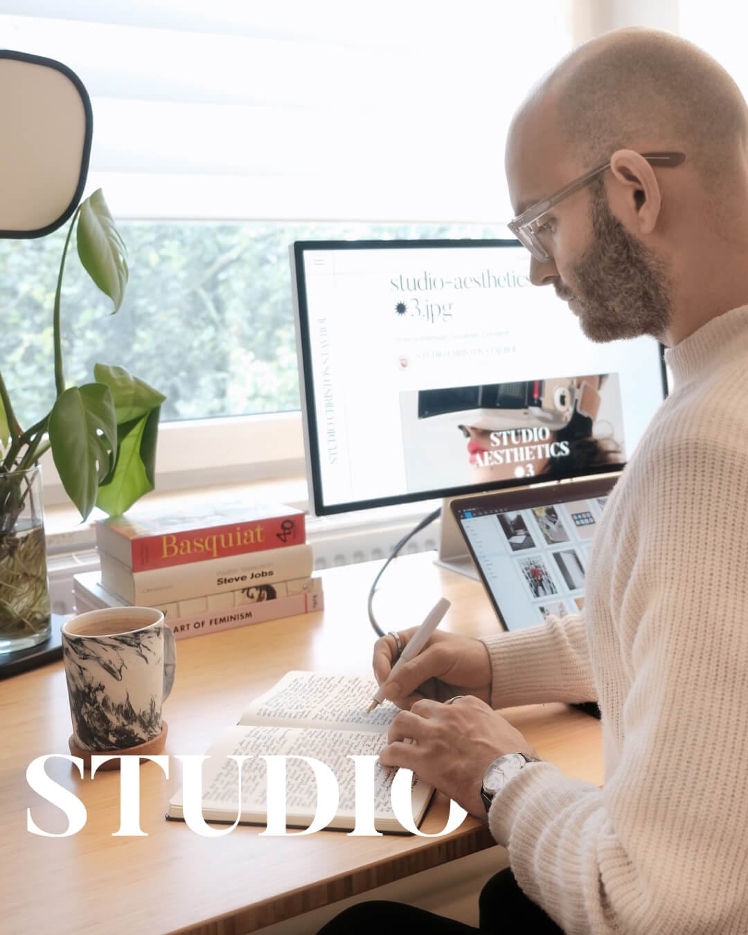 UX-Designer arbeitet am Schreibtisch mit Computern, Büchern und Pflanze. "STUDIO" im Vordergrund zeigt Fokus auf kreatives Arbeitsumfeld.